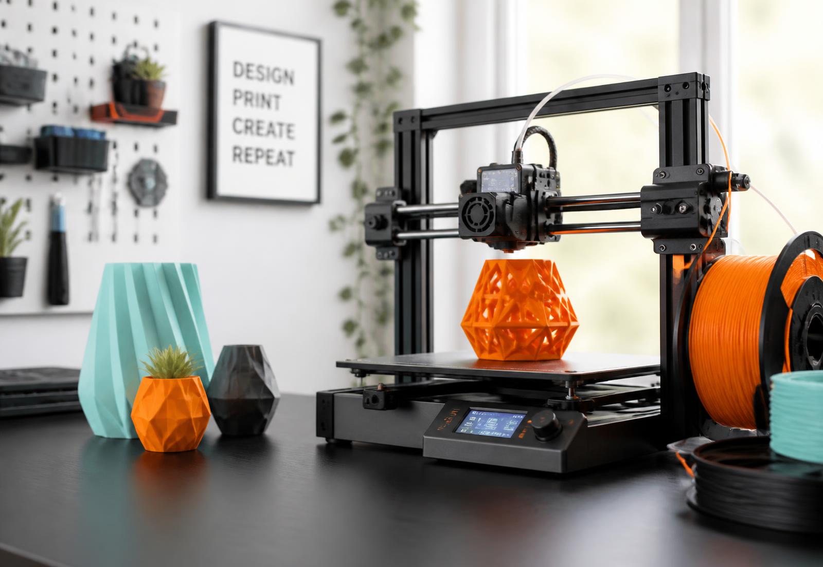 A desktop 3D printer producing an orange geometric vase beside teal and orange filament spools